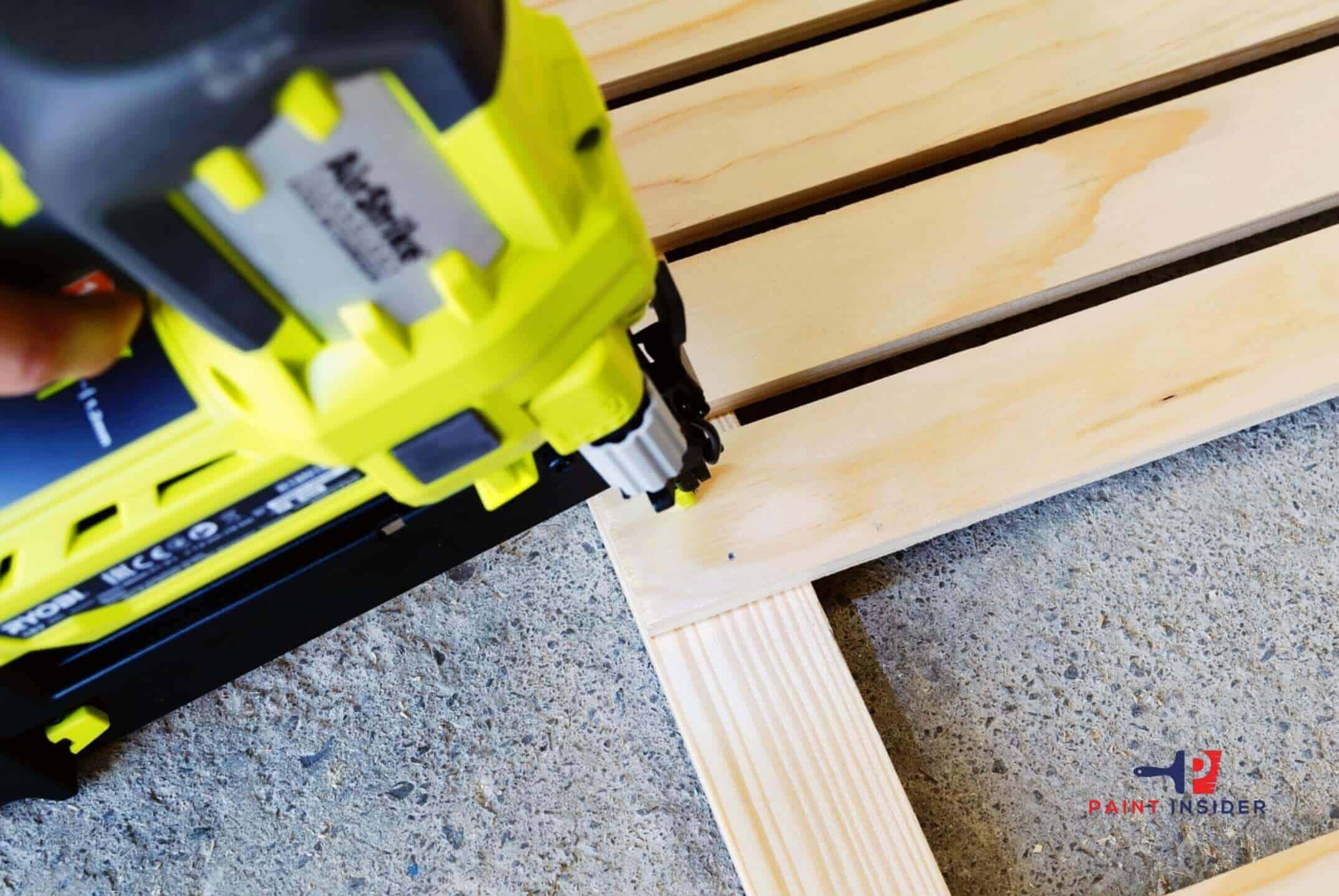 The image shows a person using a bright yellow brad nailer to secure wooden planks together on a concrete surface. The tool is being used to drive a nail into the corner where two pieces of wood meet, highlighting the precision and efficiency of the nailer. The scene illustrates a DIY woodworking or construction project. The logo of "Paint Insider" is visible in the bottom right corner of the image.